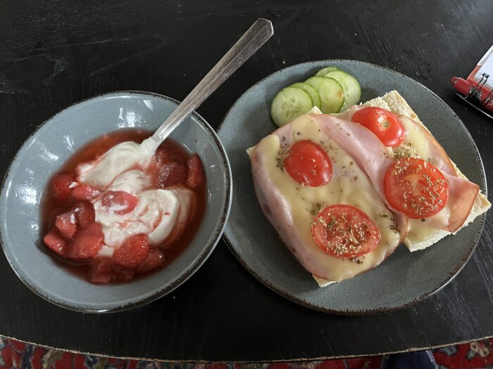 En lunch bestående av kvarg med persikor, jordgubbar och en varm baguette toppad med skinka, ost, tomater och kryddor samt skivad gurka vid sidan.