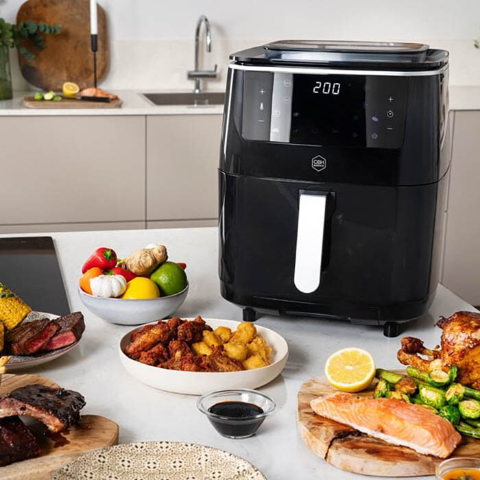 En svart Easy Fry & Grill 3in1 Steam+ airfryer står på en köksbänk, omgiven av tillagad mat, inklusive grönsaker, lax, kyckling, och friterade rätter.