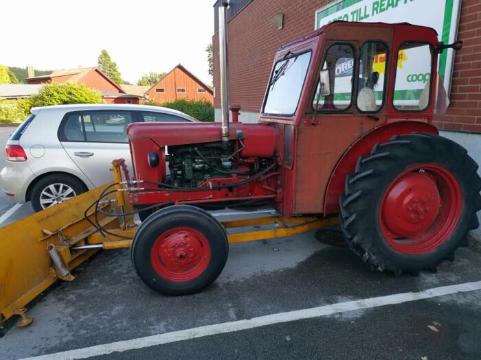 Röd traktor från 1961 med snöplog parkerad vid en vägg bredvid en silverfärgad bil.