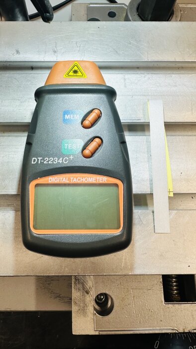 Digital tachometer DT-2234C+ placerad på en metallisk verkstadsbänk, med två orange knappar, märkt med MEM och TEST knappar samt en display.