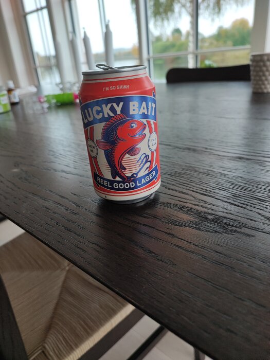 Röd burk öl med texten "Lucky Bait Reel Good Lager" på ett mörkt träbord med ett fönster och suddig bakgrund i bakgrunden.