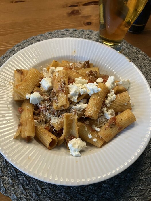 En tallrik med pasta och lammfärssås toppad med parmesan och fetaost, serverad med ett glas öl på ett bord.