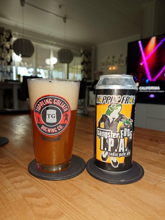 Ölglas med Troppin Goliath-bryggeri's logotyp bredvid en burk Gangster Frog IPA från Hoppin' Frog på ett bord i ett vardagsrum.