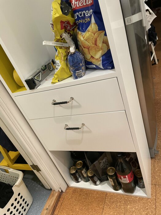 Vitt köksskåp under konstruktion med två handtag, fylld med snacks och drycker. Verktyg och verktygsmaterial synliga på hyllan.