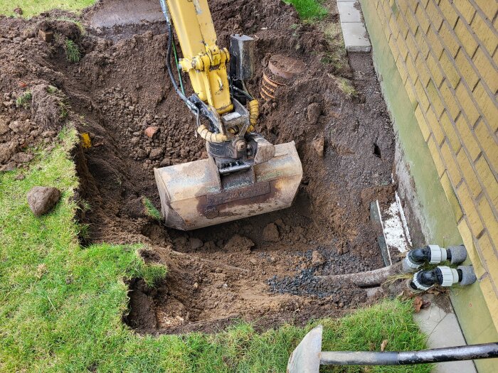 Grävmaskin i arbete vid en djup grop intill ett gult tegelhus, synliga vattenledningar och dräneringsbrunn, omgiven av gräsmatta och stenar.