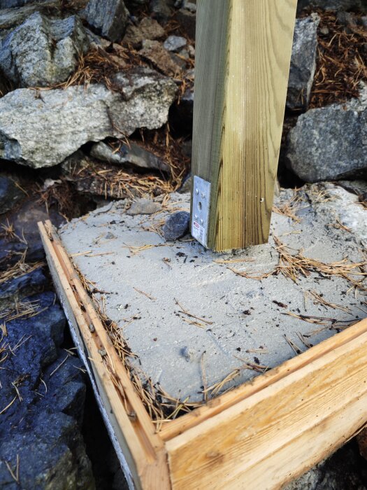 En trästolpe fäst i en betongplint bland lösa stenar, omgiven av en träform som håller betongen på plats.