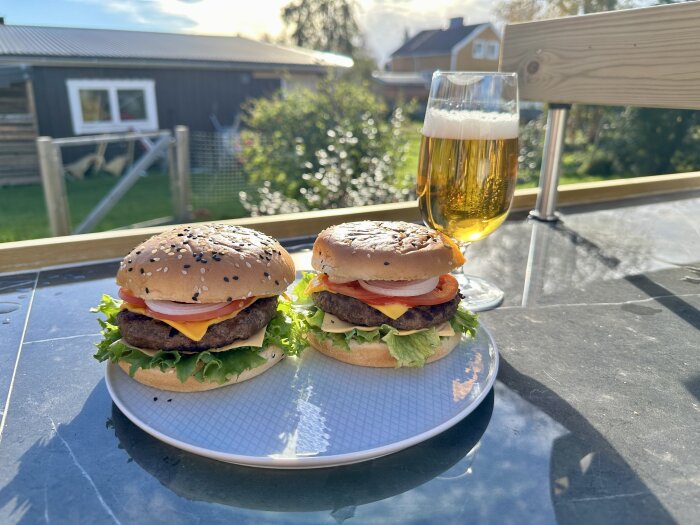 Två hamburgare på en vit tallrik och ett glas öl på ett bord utomhus, med solljus och en bakgrund av trädgård och hus.