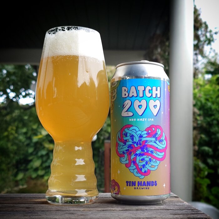 Ett fyllt ölglas bredvid en ölburk med etiketten "Batch 200 DDH Hazy IPA" från bryggeriet "Ten Hands Brewing", fotad utomhus på en regnig dag.