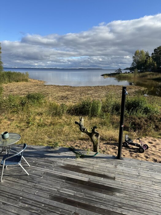 Utsikt från en altan över en sjö med lågt vattenstånd och nyklippt vass. Vid altanen står en stol och ett bord, samt en leksaksgokart på marken.