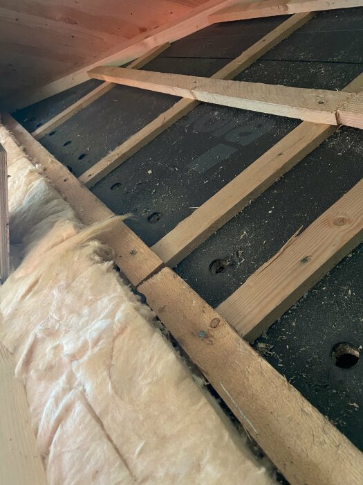 Flera borrhål i en takpanel med träreglar och isoleringsmaterial i förgrunden.