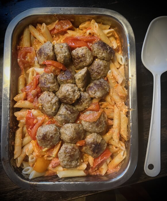 Ugnsbakad pastarätt med penne, tomater, paprika, lök och köttbullar, serverad i en rostfri ugnsform bredvid en vit serveringssked.