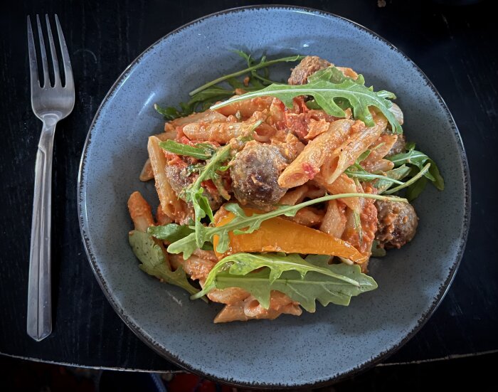 Tallrik med pennepasta, köttbullar, grillade grönsaker (tomat, paprika, lök), fetaost och ruccola. Gaffel till vänster om tallriken.