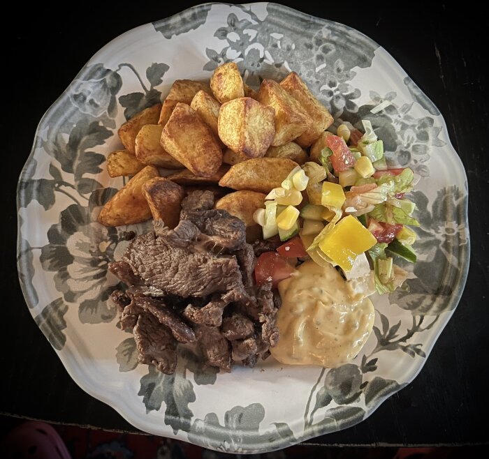 Portion med entrecote, potatisbitar med paprikakrydda, sallad och bearnaisesås på en blommig tallrik.