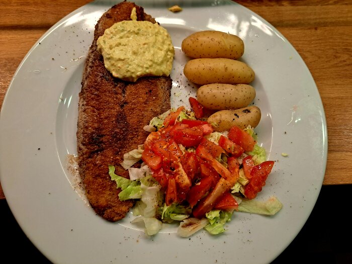 En tallrik med panerad fiskfilé toppad med senapskräm, kokt potatis, och sallad med tomater, salladsblad och dressing.