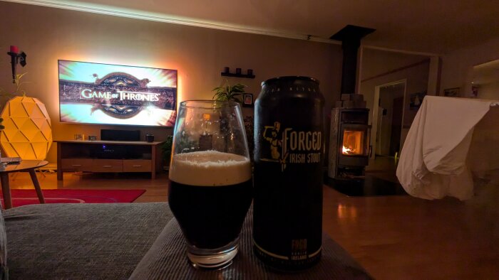 En tv visar Game of Thrones, framför står ett glas och en burk med Forged Irish Stout öl. I bakgrunden syns en tänd kamin och mysig inredning.