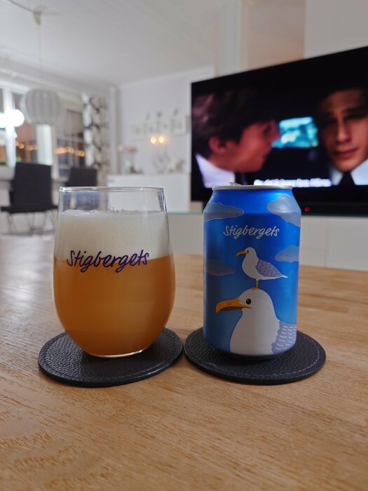 Ett glas med öl och en ölburk från Stigbergets med illustration av två måsar, placerade på ett bord med en suddig TV i bakgrunden.