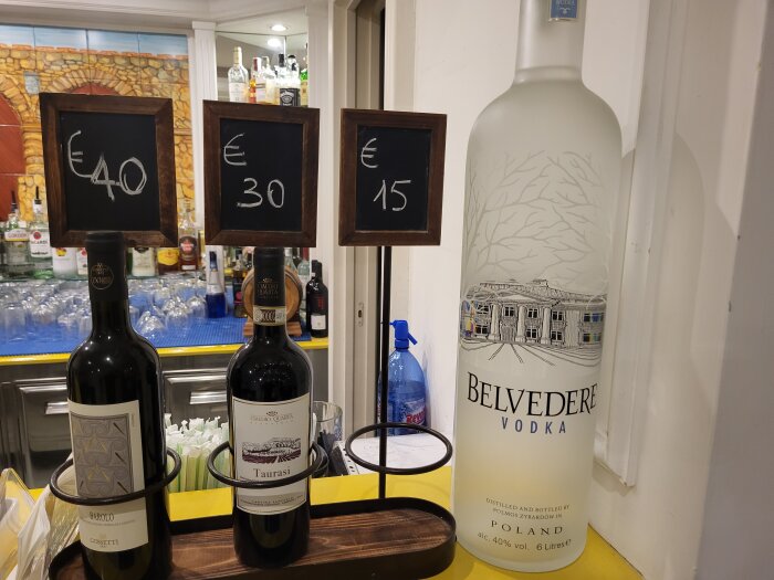 Tre flaskor sprit framför en bardisk; två flaskor rött vin märkt "Barolo" och "Taurasi" med prisskyltar på €40 och €30, samt en stor flaska Belvedere vodka.