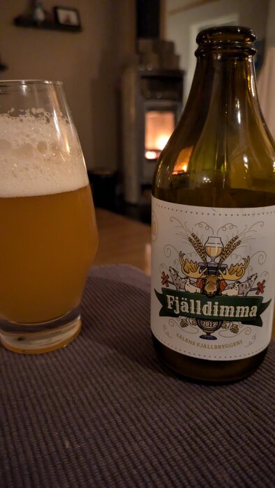 En flaska och ett glas öl med etiketten "Fjälldimma" från Sälen Fjällbryggeri, med en suddig bakgrund av en tänd kamin.