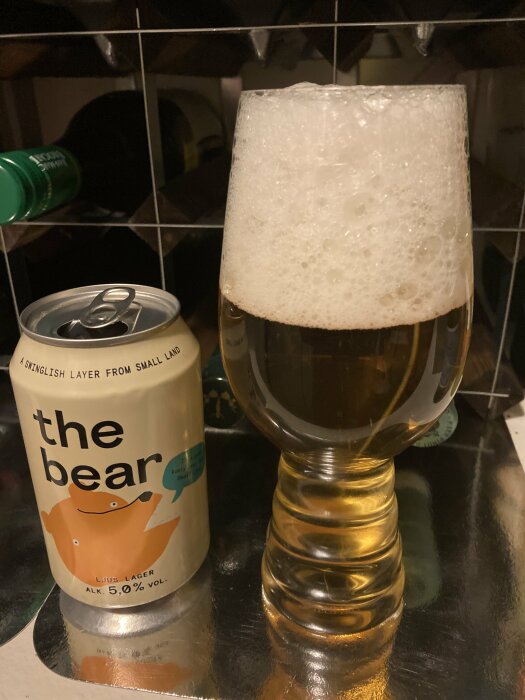 En ölburk med texten "the bear" bredvid ett ölglas fyllt med ljus öl och skum på toppen.