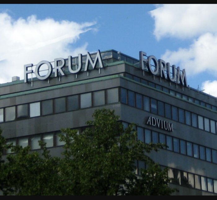 Grå byggnad med skyltar som säger "FORUM" på taket och "ADVIUM" på fasaden, med träd och en blå himmel i bakgrunden.