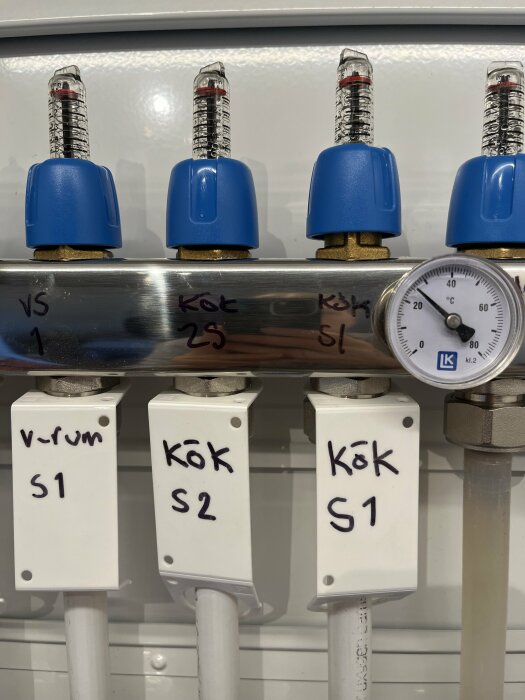 Fyra flödesmätare med blåa toppar och en temperaturmätare på ett rörsystem för golvvärme. Etiketter visar "Vrum S1", "Kök S2", "Kök S1".