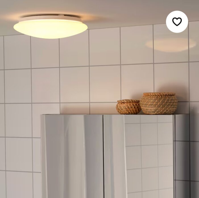 Vit rund LED-taklampa från IKEA-serien Barlast, monterad i ett rum med vita kaklade väggar och brynflätade korgar ovanpå ett skåp.