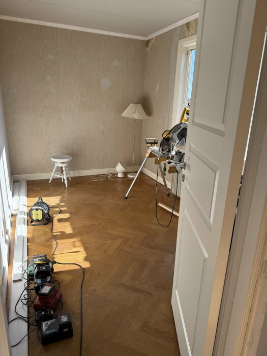 Rum under renovering med pågående spackling, arbetslampor, verktyg och material ligger på trägolvet, och dörr delvis öppen.