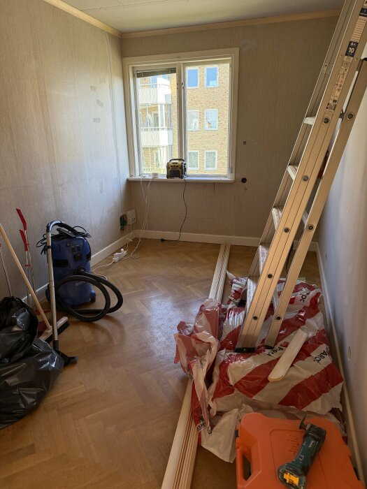 Ett rum under renovering med en stege, verktyg och byggmaterial på golvet. Spackling pågår och ett fönster syns i bakgrunden.