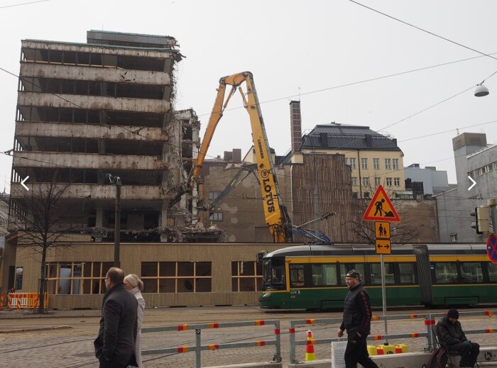 En byggnad under rivning med en stor grävmaskin, en spårvagn passerar i förgrunden och personer går förbi på gatan.