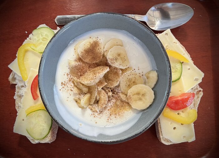 En skål yoghurt toppad med bananskivor, kanel och müsli, omgiven av två smörgåsar med ost, gurka, paprika och tomat.