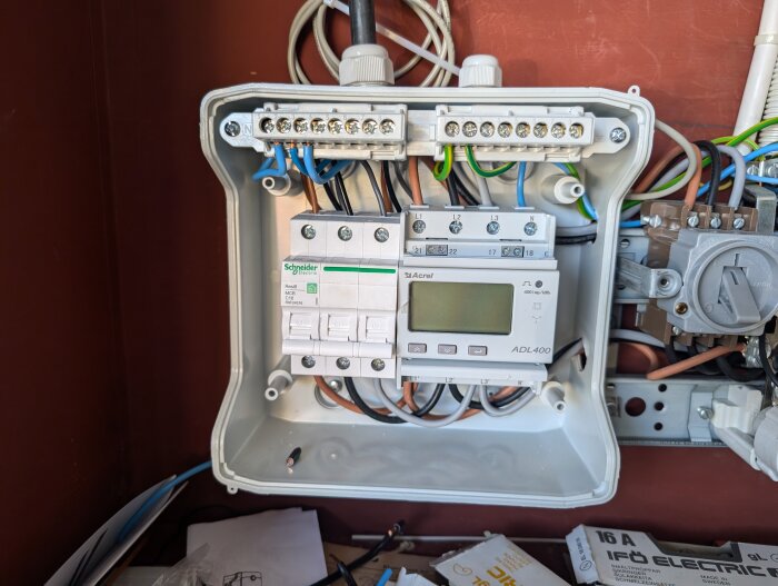 Elcentral med en Schneider Electric mätare och Acrel lastbalanserare monterade på en brun vägg, omgivna av kablar och ledningar.