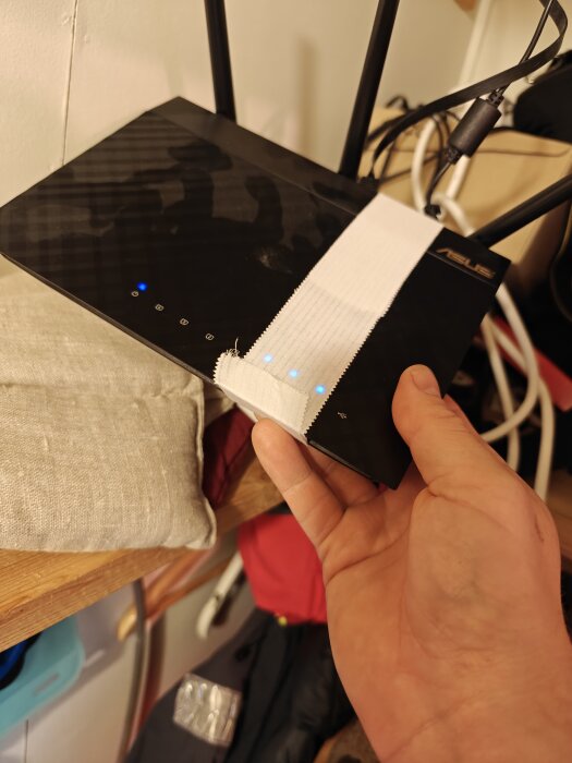 En hand håller en trådlös router som är tejpad för att hålla on/off-knappen intryckt, vilket får routern att lysa med blå indikatorer.