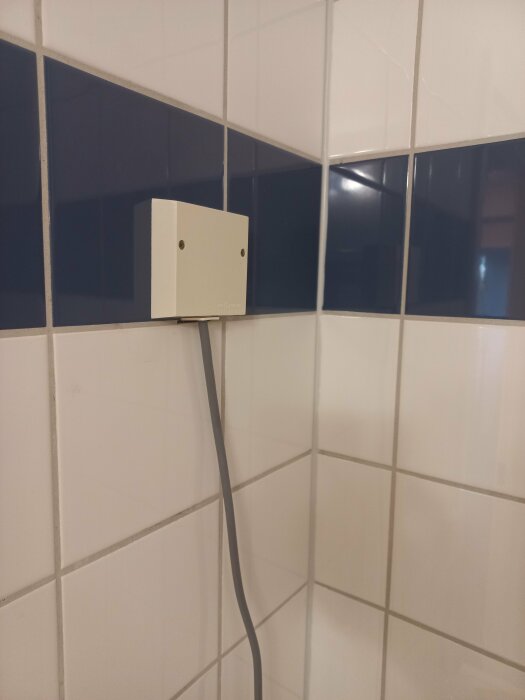 Elkabel insatt i en vit dosa fastsatt på en kaklad vägg i ett badrum, eventuellt för elinstallation över tvättmaskin.