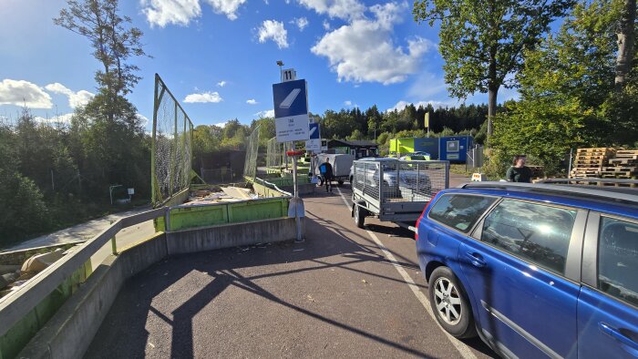 En återvinningsstation med containrar för träavfall, där en parkerad blå bil med släp som är under lossning syns i förgrunden och skog i bakgrunden.