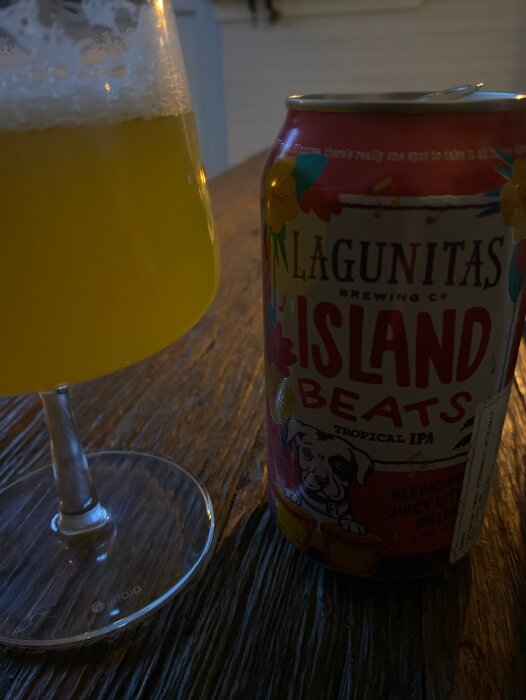 Burk med Lagunitas Island Beats Tropical IPA och ett glas fruktig öl på ett träbord i svagt upplyst miljö.