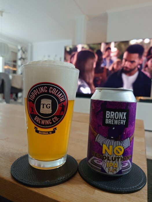 Ett glas med öl från Toppling Goliath Brewing Co. och en ölburk från Bronx Brewery, "No Resolutions IPA", står på ett bord framför en TV.