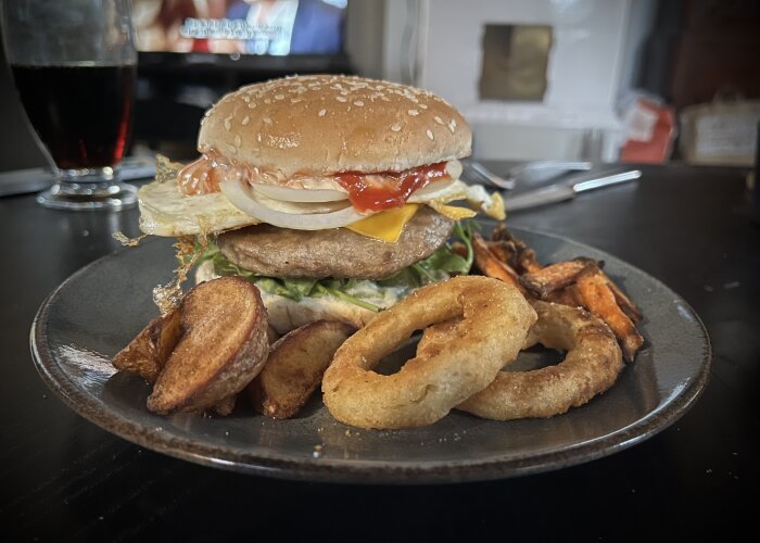 En hamburgare med skivad lök, tomat, ägg, ost, ruccola, senap och ketchup. På tallriken finns också klyftpotatis, sötpotatispommes och lökringar.