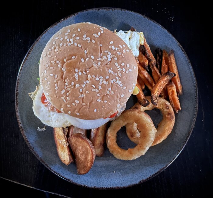 En tallrik med en hamburgare med sesambröd, klyftpotatis, sötpotatispommes och lökringar. Burgaren har tomat, sallad och vad som ser ut som ett stekt ägg.