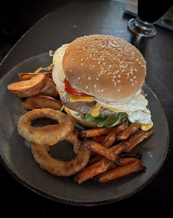 Tallrik med en hamburgare som innehåller tomat, ägg, ost, ruccola, senap, ketchup och skivad lök, samt klyftpotatis, sötpotatispommes och lökringar.