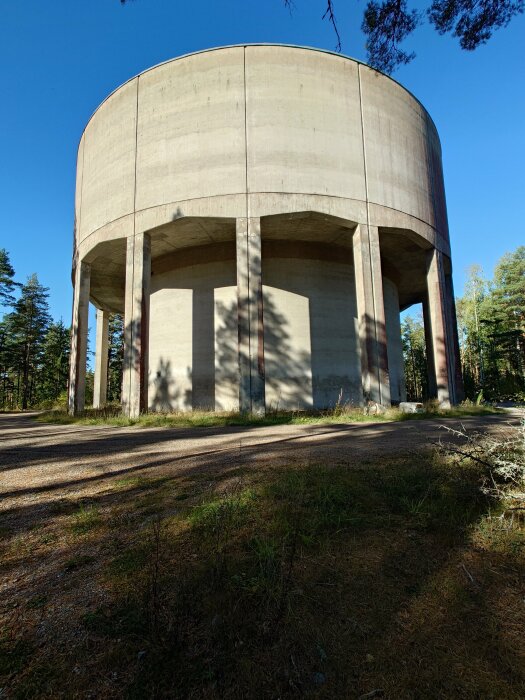 Betongvattenreservoar med höga pelare, omgiven av skog under en klarblå himmel.