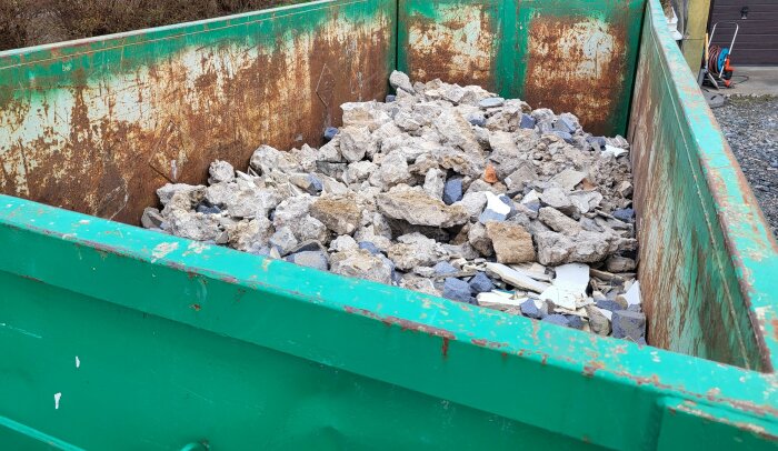 En grön container fylld med blandade betong- och väggrester från en byggarbetsplats. Containern är sliten och rostad på insidan.