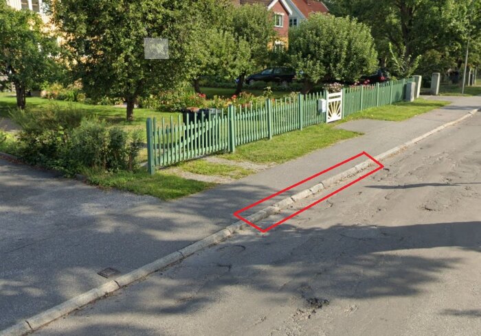 Trottoar och gata med en fyra meter lång sträcka av kantsten markerad i rött. Grön staket och hus med trädgård i bakgrunden.