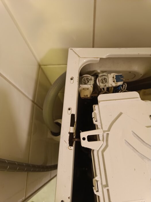 Öppen tvättmaskin med synliga fjädrar och komponenter, fotad ovanifrån. Väggar med kakel och en slang syns i bakgrunden.