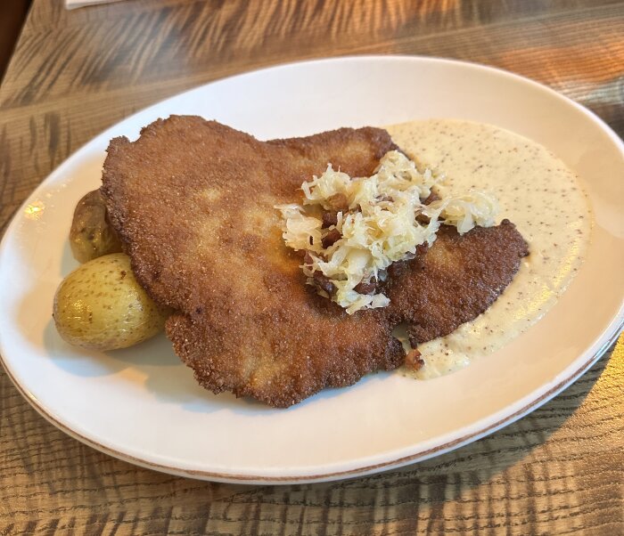 En portion schnitzel med potatis, surkål och sås på ett vitt fat, serverad på ett träbord.