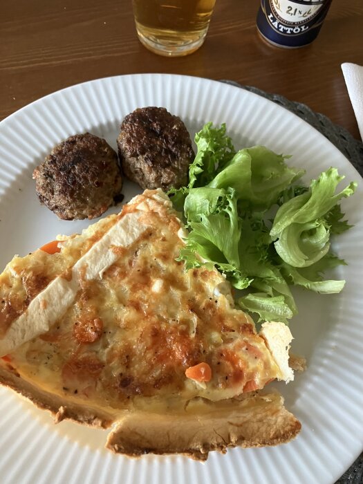 En tallrik med en bit paj med grönsaker, två köttbullar och en liten portion grönsallad. I bakgrunden syns ett glas och en burk lättöl.