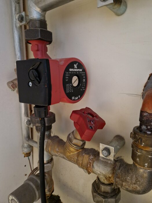 Närbild på en röd Grundfos vattenpump och rörkopplingar kopplade till en gaspanna, med en svart kabel ansluten till pumpen.