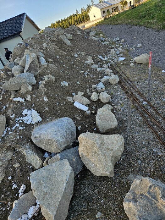 En hög med fyllnadsmassor och stenblock på en grusväg, med hus och grönområde i bakgrunden i Gemla, Växjö Kommun.