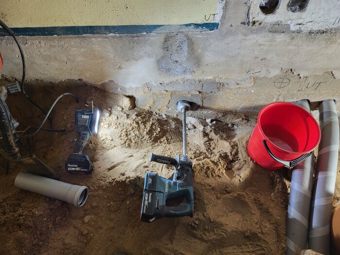 Makita borrmaskin, röd hink och ventilationsrör på mark fylld med sand framför en vägg.