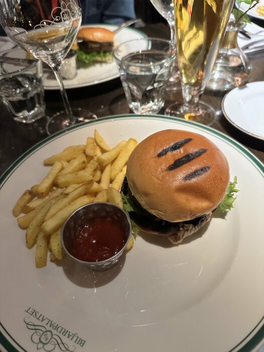 Tallrik med hamburgare, pommes frites och ketchup på en restaurang. Ett glas vatten och en ölglas syns i bakgrunden.