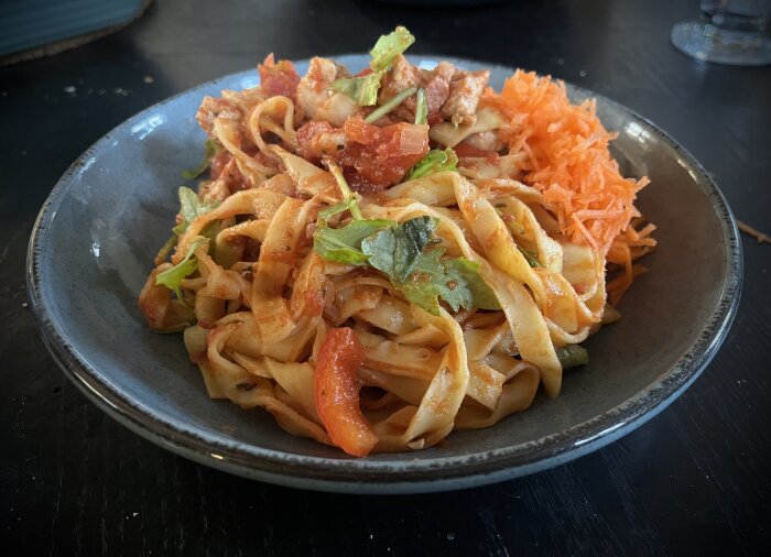 Pasta i tomatsås med kyckling, fläskbitar, ruccola samt riven morot på en grå tallrik på ett mörkt bord.